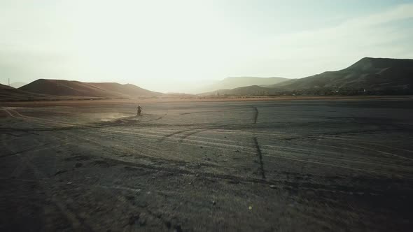 Drone View of Man on Motorbike Rides on a Dry Lake with Dust and Beautiful Hills on Background in