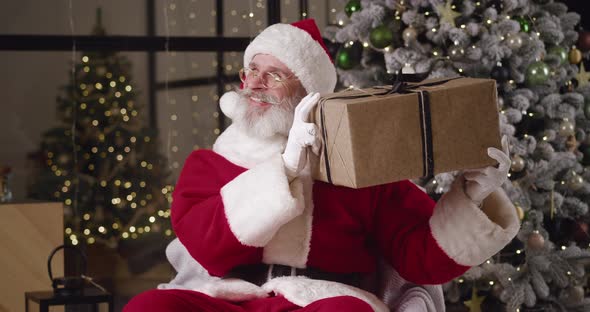 Kind Mature Man with Gray Beard Dressed As Santa Claus Sits on Armchair Against Decorated Christmas alt