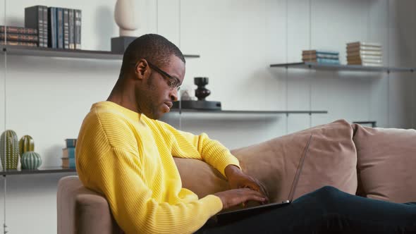 Young guy working on a laptop on the couch. Young man typing with laptop on couch at home alt