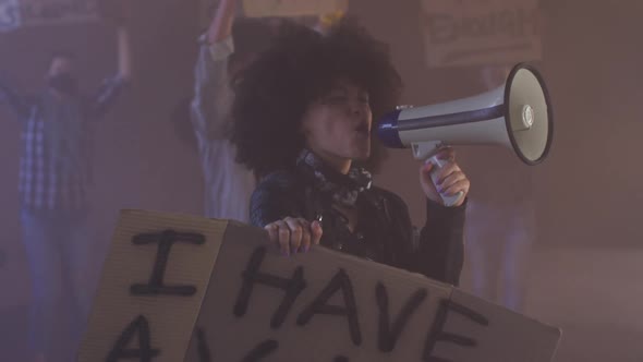 Diverse group of people wearing face masks holding protest signs shouting into megaphone alt