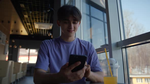a Young Guy is Sitting in a Cafe By the Big Window Drinking Juice and Having Fun on the Internet