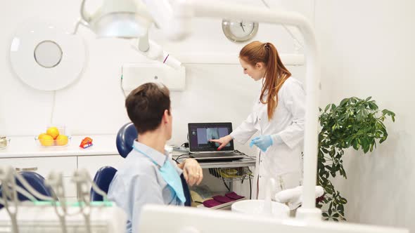 Young Female Dentist Pointing at Xray and Recommending Dental Treatments to Male Patient alt