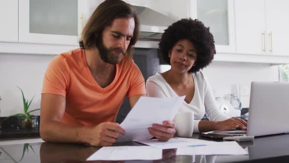 Mixed race couple using laptop and calculating finances in the kitchen at home alt