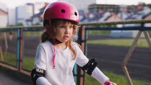 Cute Light Hair Little Girl with Big Eyes in Pink Helmet in Elbow and Knee Pads Rides a Bicycle