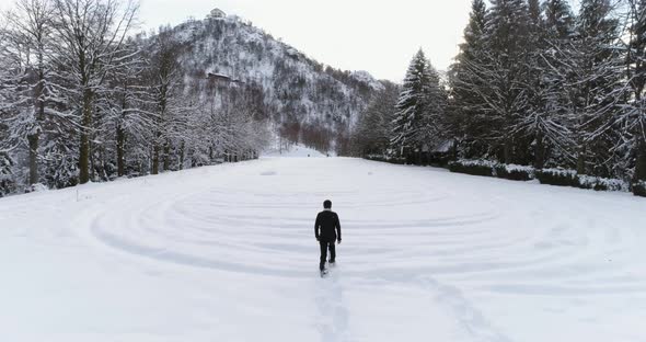 Forward Aerial Over Man Walking with Snowshoes on Mountain Snow Covered Field Near Pine Forest Woods alt