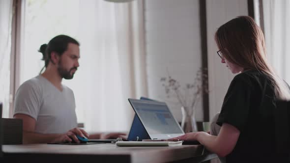 A young man and a young girl are working in front of laptops in their bright apartment. alt