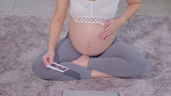 Top view Pregnant Woman sitting on carpet using computer laptop and holding ultrasound image alt