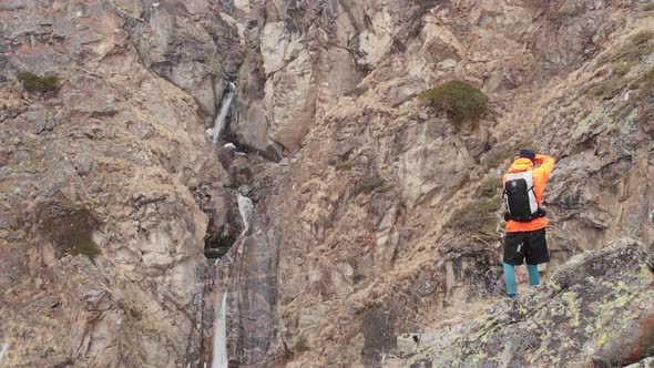Aerial View of a Young Male Photographer with a Camera in His Hands Stands on a High Rock in a Gorge alt