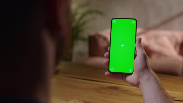 Handheld Camera Back View of Young Man at Home Sitting on a Wood Desk Using With Green Mockup Screen alt