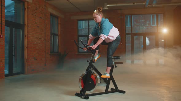 Athletic Woman Riding on Spinning Stationary Bike Training Routine in Haze Gym Weight Loss Indoors alt