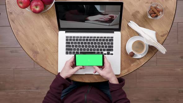 Man in Coffee Shop Watches a Video and Holds the Smartphone with Green Screen on alt