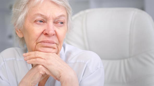Thoughtful grey haired senior lady looks into window holding head over hands alt