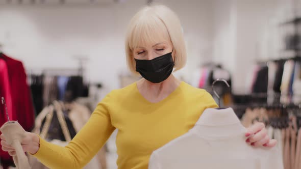 Mature Elegant Woman Shopper in Medical Mask Stands in Clothing Store Chooses Clothes Holds Two alt