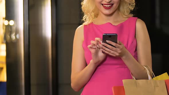 Girl with Shopping Bags Holding Cellphone and Scrolling Screen Online Discounts alt