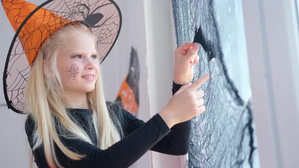 Happy Girl in Witch Costume Preparing for Halloween Decorating Window in Room By Black Bats alt