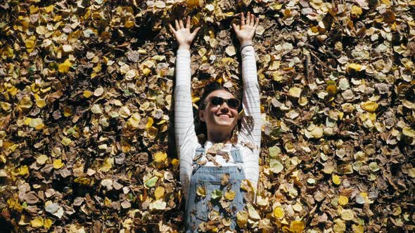 Cheerful Young Woman in Denim Overall is Lying on Autumnal Yellow Leaves