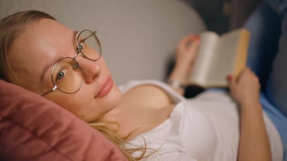 Caucasian Woman Holding Book Lying on Sofa and Looking at you at Home alt
