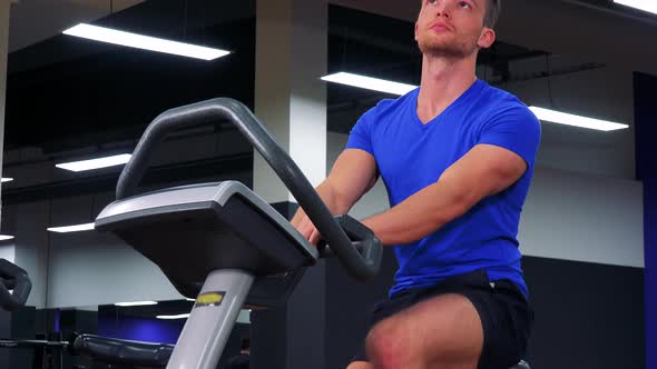A Young Fit Man Trains on an Exercise Bike in a Gym - Closeup From Below alt