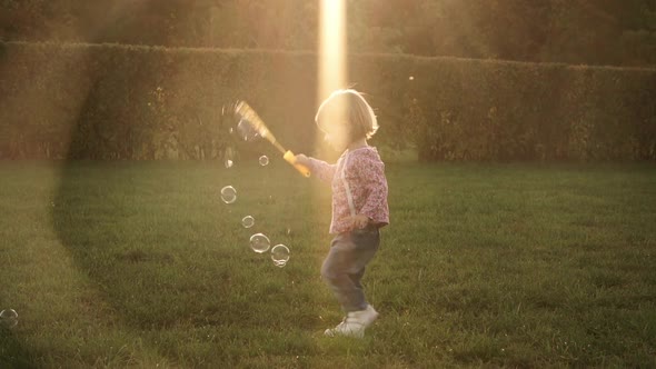 Little Baby Girl Blows Soap Bubbles on Green Lawn in Summer Park at Sunset Outdoors