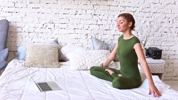 A Slender Woman in a Green Bodysuit Performs Yoga Exercises Online Sitting on a White Bed