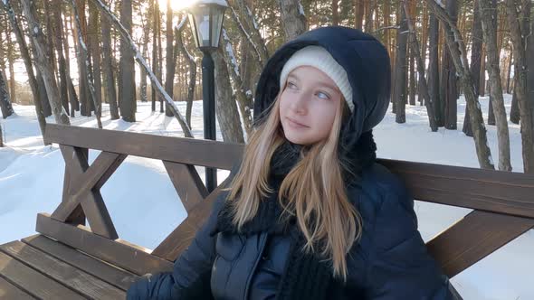 Thoughtful Pretty Blonde Girl in Black Coat Sitting on Wooden Bench in Winter Snowy Park Enjoying alt