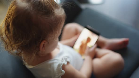 Smiling Little Girl is Sitting on Armchair and Using Smart Phone alt