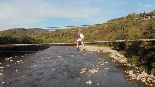 Aerial Drone View of Young Man in White Shirt Sitting on Wooden Bridge Over Mountains River and