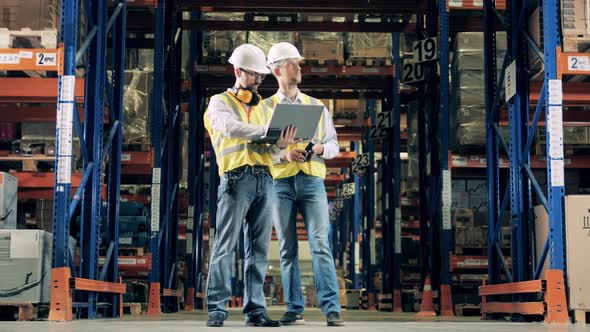 Two Warehouse Workers in Uniform and Helmets Discussing Business Issues alt