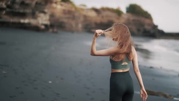 Backside View Woman in Sportswear Walks Along Black Sand Beach and Turns Around alt