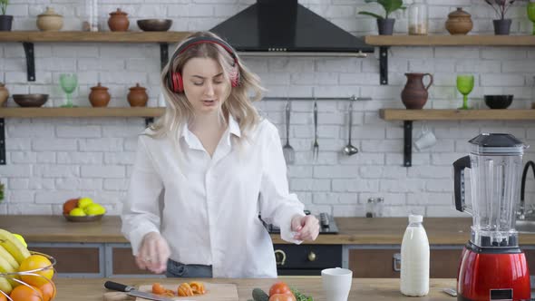 Portrait of Cheerful Beautiful Young Woman in Headphones Dancing and Singing As Cooking Healthful alt