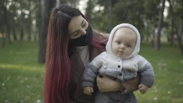 Happy Young Millennial Mother on Covid19 Face Mask Carrying Cute Infant on Hands alt