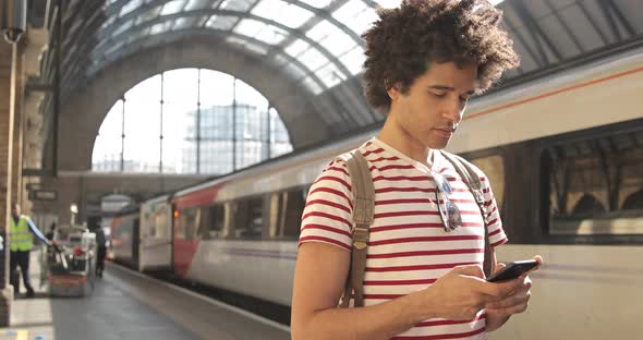Man at train station checking timetables on mobile phone alt