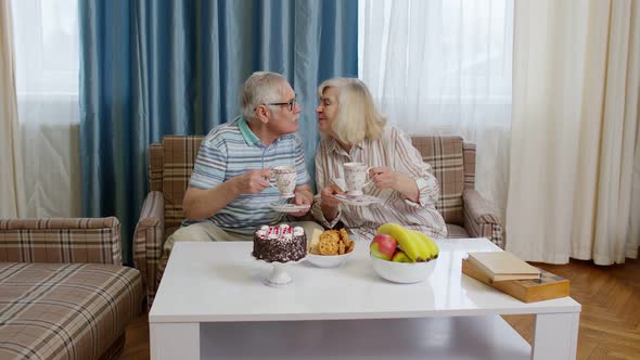 Senior Couple Holding Drinking Tea Talking Laughing Smiling Kissing Sitting on Sofa at Home alt