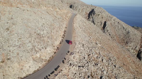 Aerial drone shot of Red car driving along the narrow coastal road above rocky shore alt