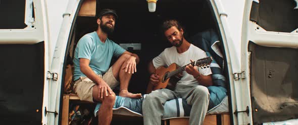 Two men sitting in back of a van singing and playing the guitar alt