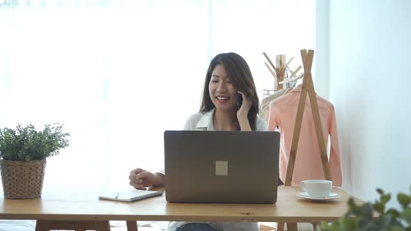 young smiling asian woman working on laptop while sitting in a living room using phone. alt