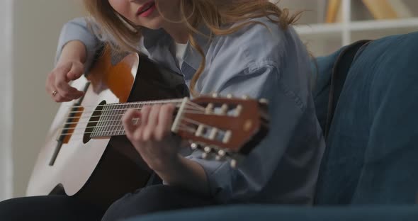 Young Female Musician Is Plucking Strings of Guitar Sitting on Couch in House Lover of Country Music alt