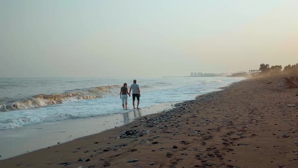 A Couple a Man and a Woman Walk Hand in Hand Along a Sandy Beach Barefoot on the Incoming Waves at