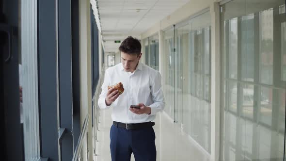 Young Man Types on the Mobile Phone and Eats Burger in Corridor of Office Centre alt