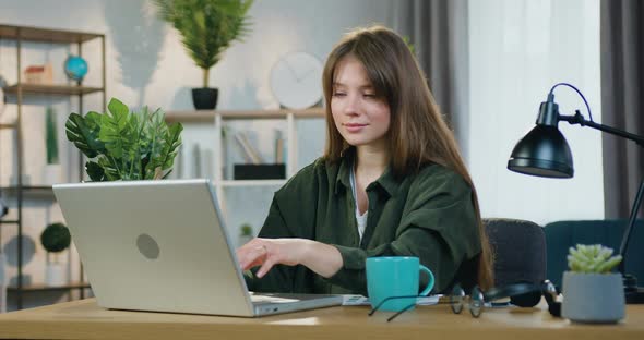 Young Girl in Casual Clothes Sitting at Her Workplace in Home Office and Working on Computer alt