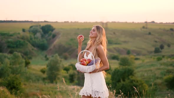 Beautiful sexy blonde girl in white dress posing in a field at sunset with a basket of fruit