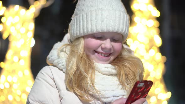 Beautiful Blonde Woman Chatting in a Smartphone on a Christmas Decorated Street in Winter