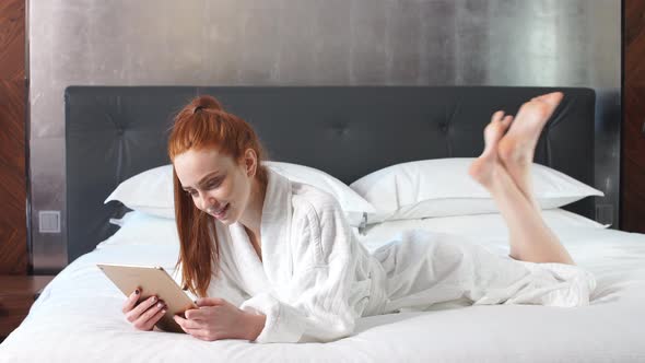 Young Redhead Woman Using Tablet Computer Lying on Bed in Hotel Room.