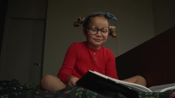 Little Girl in Glasses Lying on the Bed and Reading Book alt
