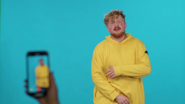 Young Guy in Yellow Hoodie Dances and Singing to the Music on a Blue Background and Takes a Selfie alt