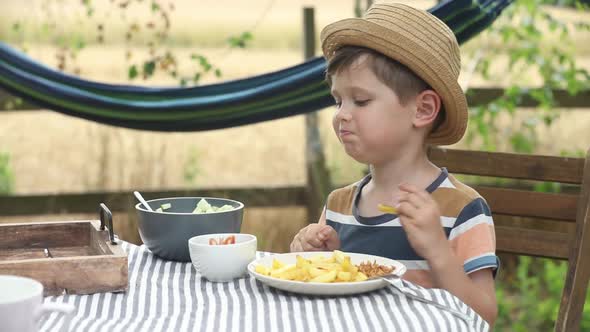 Little boy sit at table in a garden and eating  salad and french fries