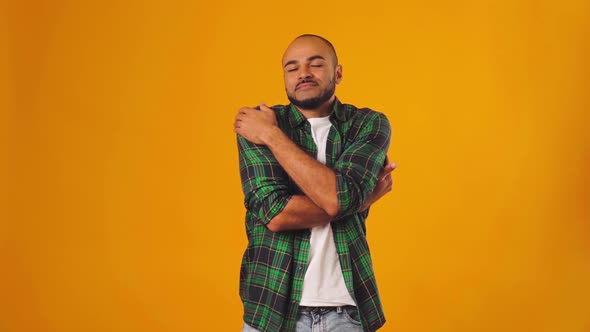 Young African American Man Hugging Himself and Feeling Comfortable Against Yellow Background alt