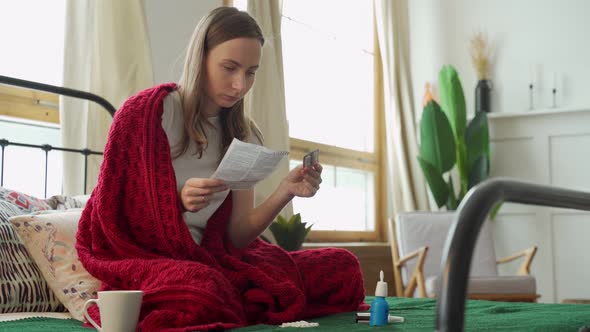 Woman Reads the Instructions of the Pill Sitting on the Bed Covered with a Blanket alt