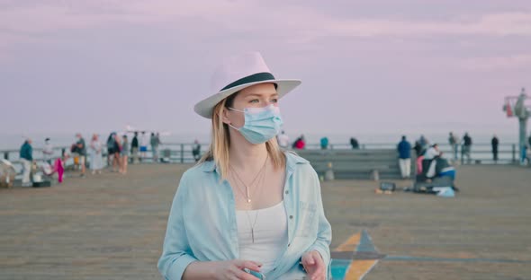 Portrait of Traveler Woman in COVID-19 Mask Enjoying Pink Sunset on Ocean Pier alt