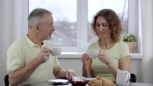 An Elderly Couple is Having Breakfast and Talking at a Table By the Window